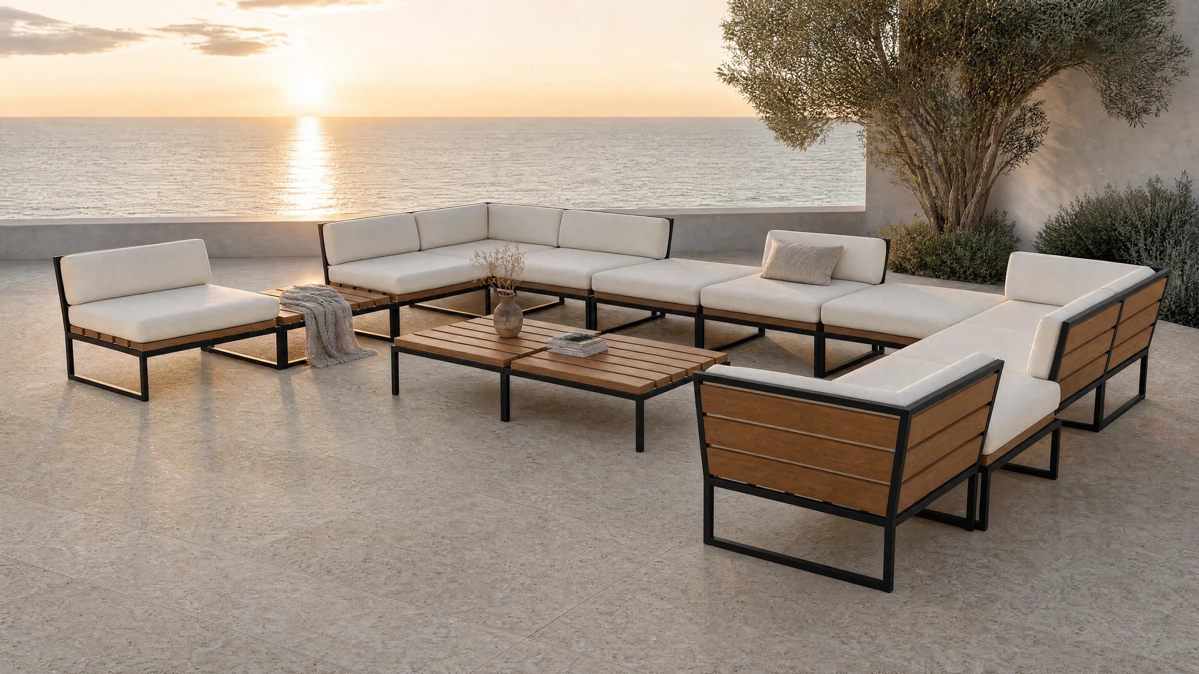 A Hawa arrangement on a clifftop terrace at sunset: separate 800 millimetre modules on thin black tube frames lining three sides of an open centre, wide timber slats showing under thick loose cushions, slatted backs leaning away with plump pillows against them, and two low slatted tables side by side in the middle, with the low sun over the sea beyond.