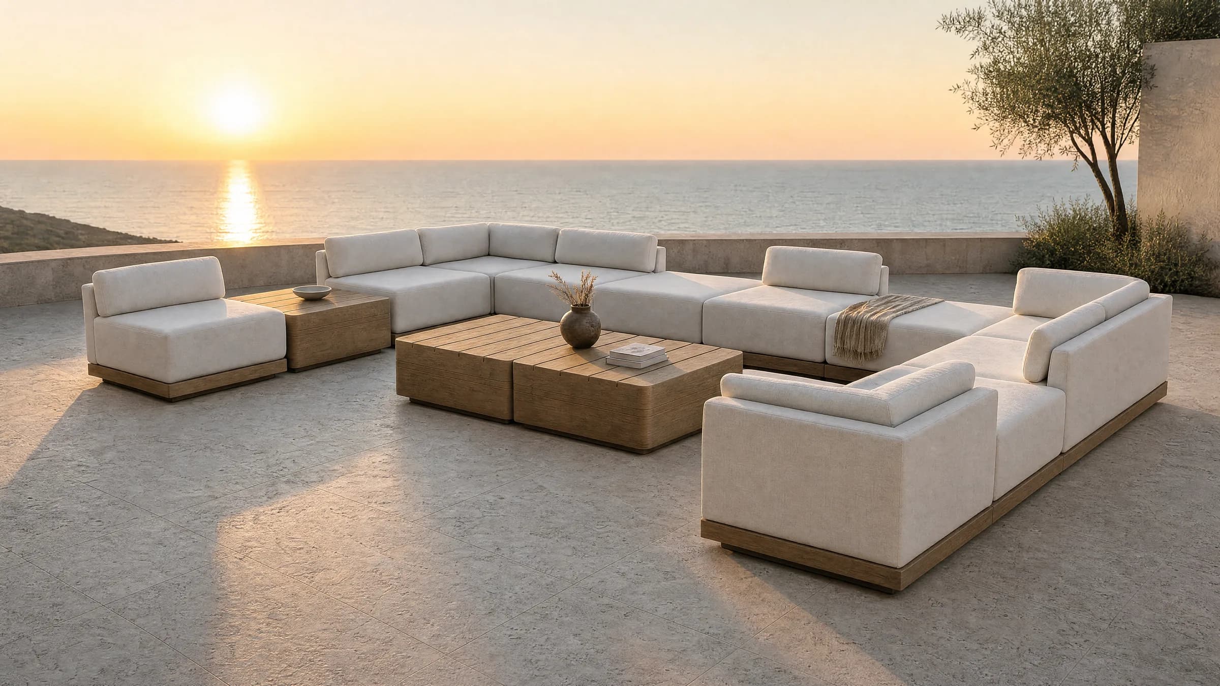 A Rima arrangement on a clifftop terrace at sunset: separate 800 millimetre upholstered modules lining three sides of an open centre, each soft block standing on a slim timber base, loose cushions leaning against the upholstered backs, and two low timber tables side by side in the middle, with the low sun over the sea beyond.