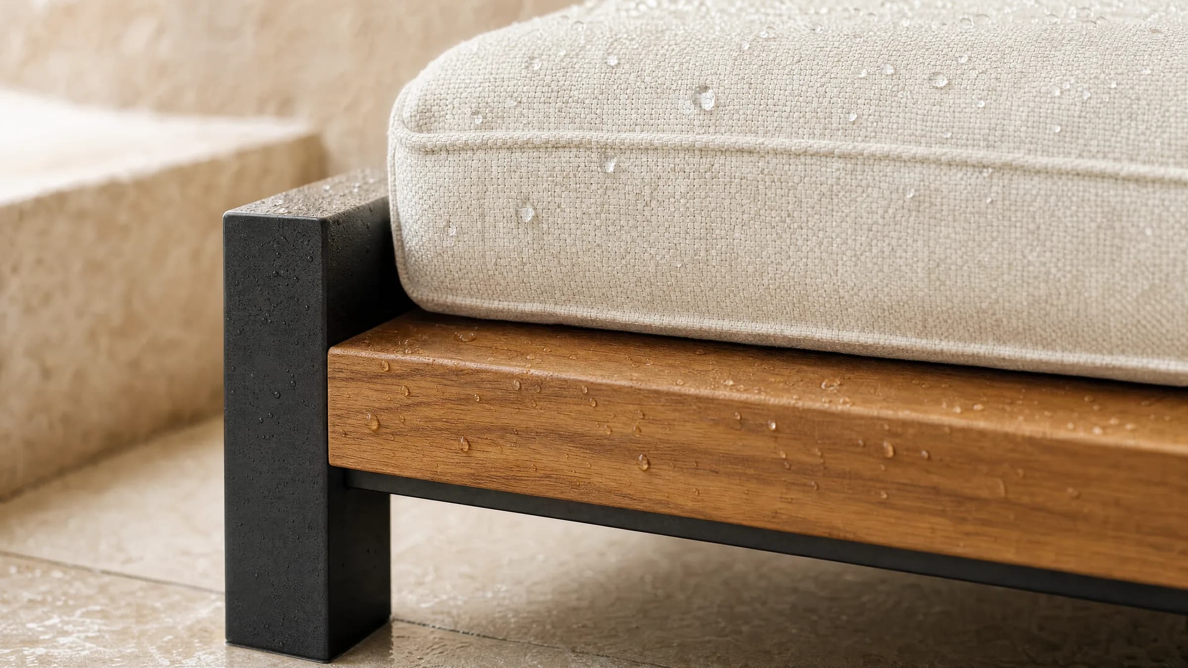 A close view of the base corner of an outdoor seating module after a rinse: a rail of warm oiled teak runs along the base outside the cushion, meeting a matte black powder-coated frame at a right angle, with fresh water beaded on both the timber and the edge of the off-white woven cushion above.