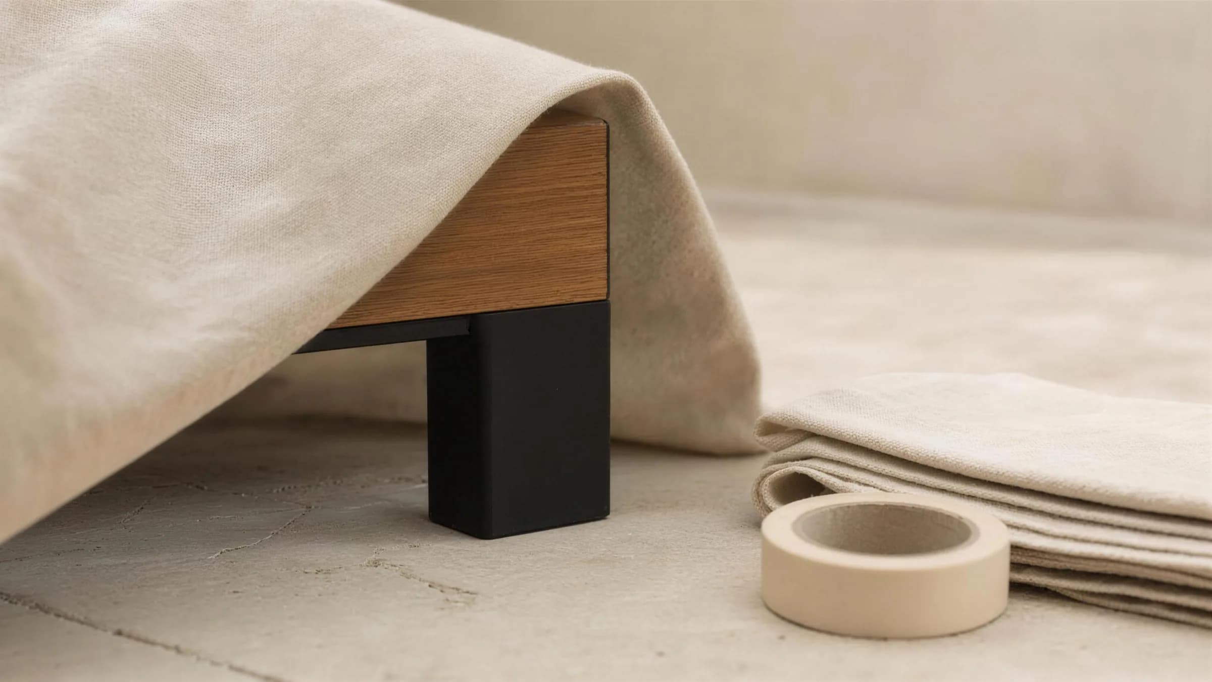 A delivery, part unwrapped, on a pale stone terrace: a loose natural cotton dust sheet folded back from one corner of a piece to show a matte black powder-coated leg and a rail of oiled teak, with a neatly folded stack of the same cloth and a roll of plain tape on the floor beside it.