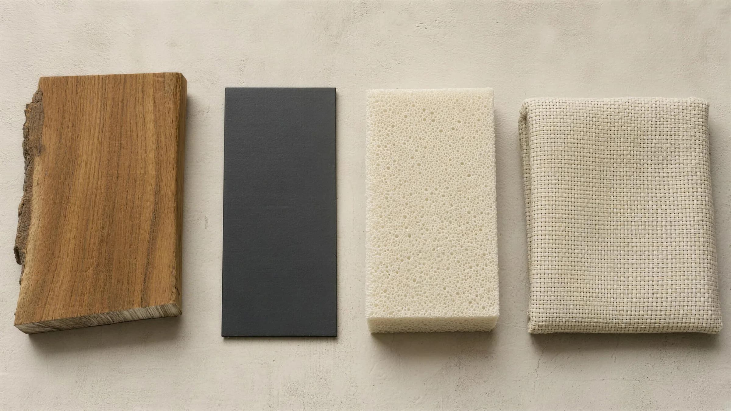 Four material samples laid out in a row on pale limewashed plaster and photographed from above: a sawn offcut of warm oiled teak with the grain and a live edge showing, a flat chip of matte black powder-coated metal, a cut block of pale open-cell quick-dry foam, and a folded square of warm off-white solution-dyed acrylic weave.
