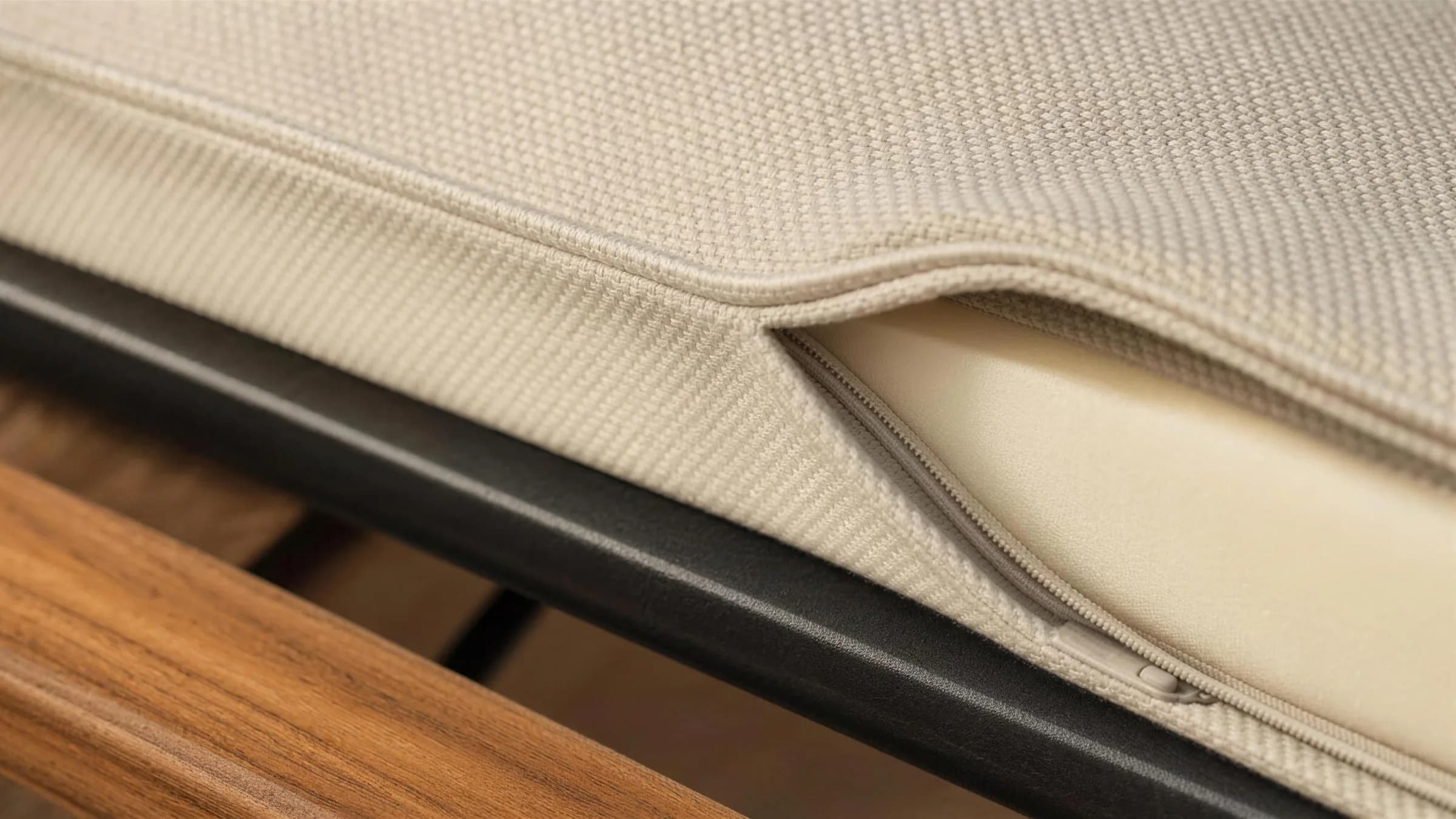 A close view of a seat cushion with its cover drawn back at one corner, showing the piped seam, the stitching, the concealed zip and the pale foam core inside, with the matte black frame and oiled teak of the module below.