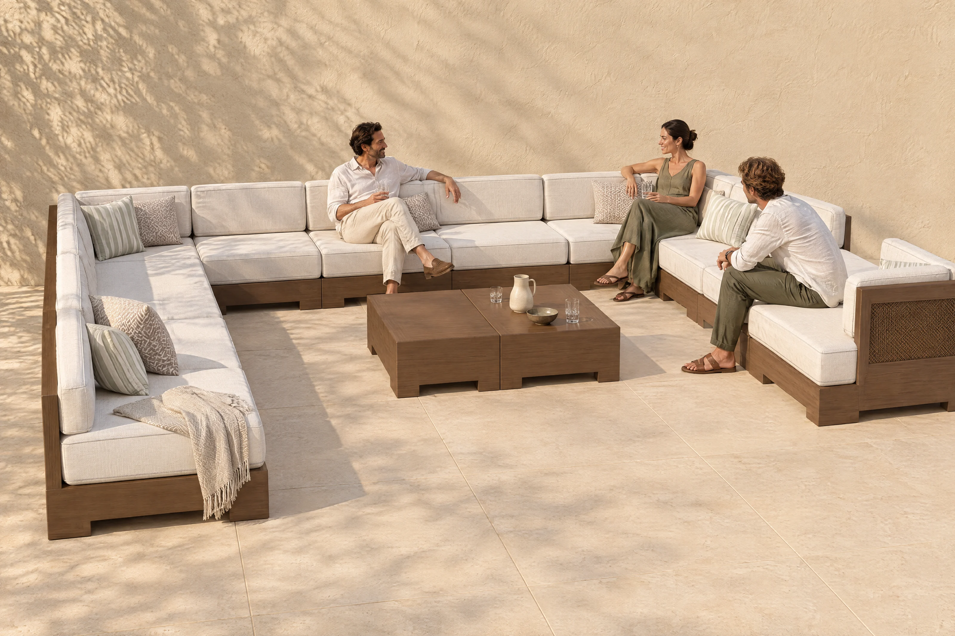 A Nasim arrangement in the founder's own U on a limestone terrace, Bliss Linen cloth on walnut-coloured timber with matching woven cane panels, three people talking, the shadow of an olive tree on the plaster wall.