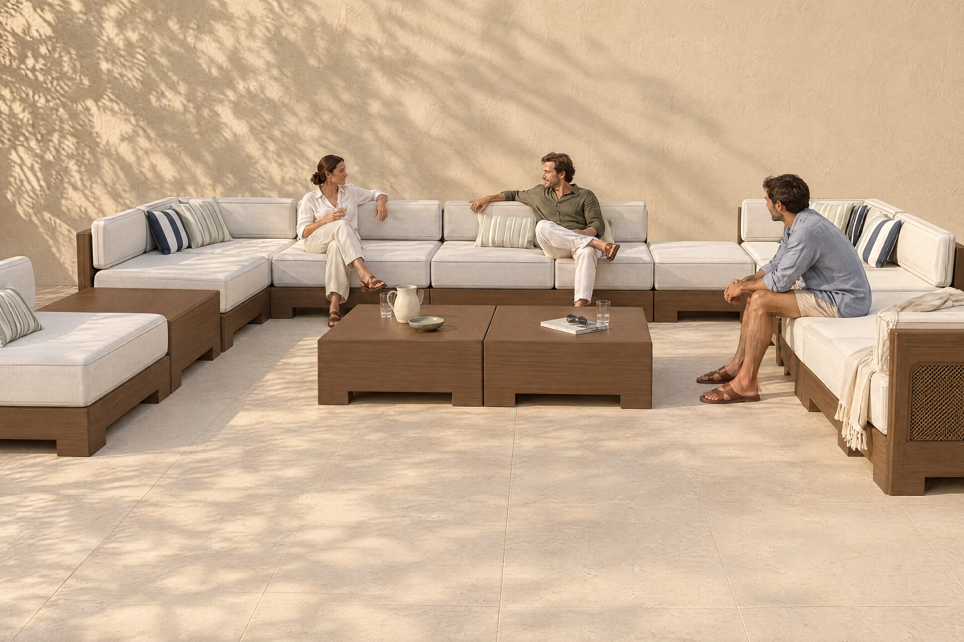 A Nasim arrangement in the founder's own U on a limestone terrace, Bliss Linen cloth on walnut-coloured timber with matching woven cane panels, three people talking, the shadow of an olive tree on the plaster wall.