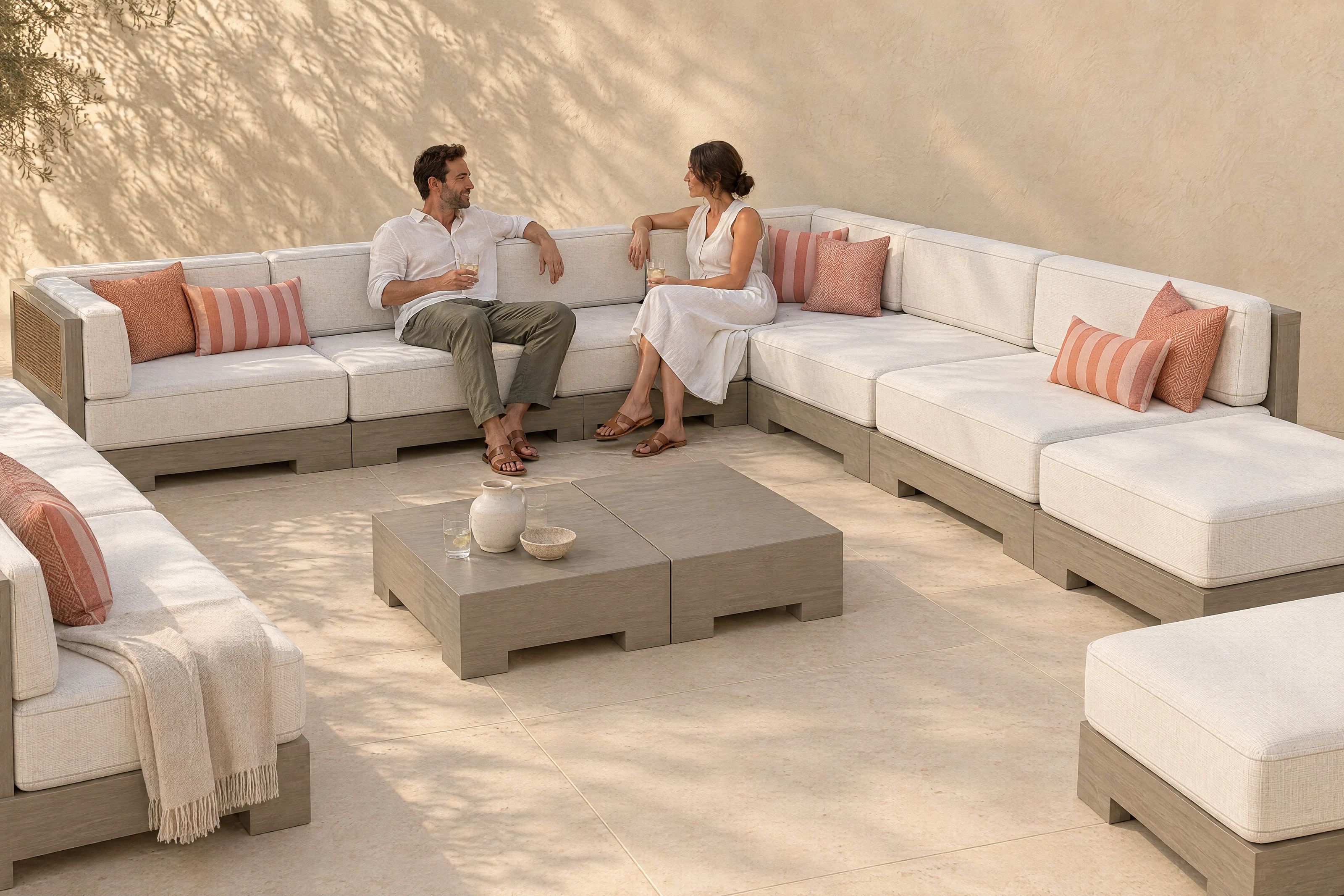 A Nasim arrangement in the founder's own U on a limestone terrace, Bliss Linen cloth on aged grey timber with matching woven cane panels, a couple talking, the shadow of an olive tree on the plaster wall.