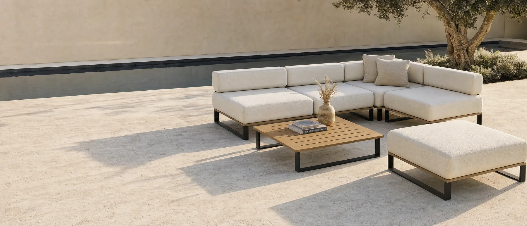 A Nour arrangement in a walled courtyard in flat daylight: a long run against the wall with a corner returning at one end, a low timber table in front of it and an ottoman set apart, on pale stone paving.