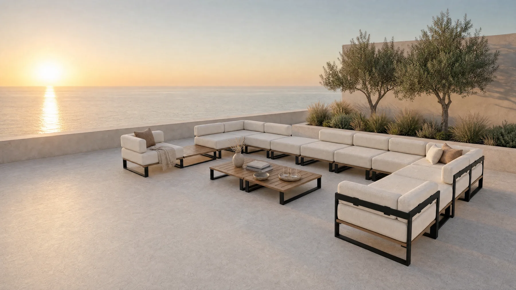 A Nour arrangement on a stone terrace at sunset above the sea: a long run with a corner returning at each end, two low tables inside it and an ottoman set apart, an olive tree at the parapet and the sun low on the water.