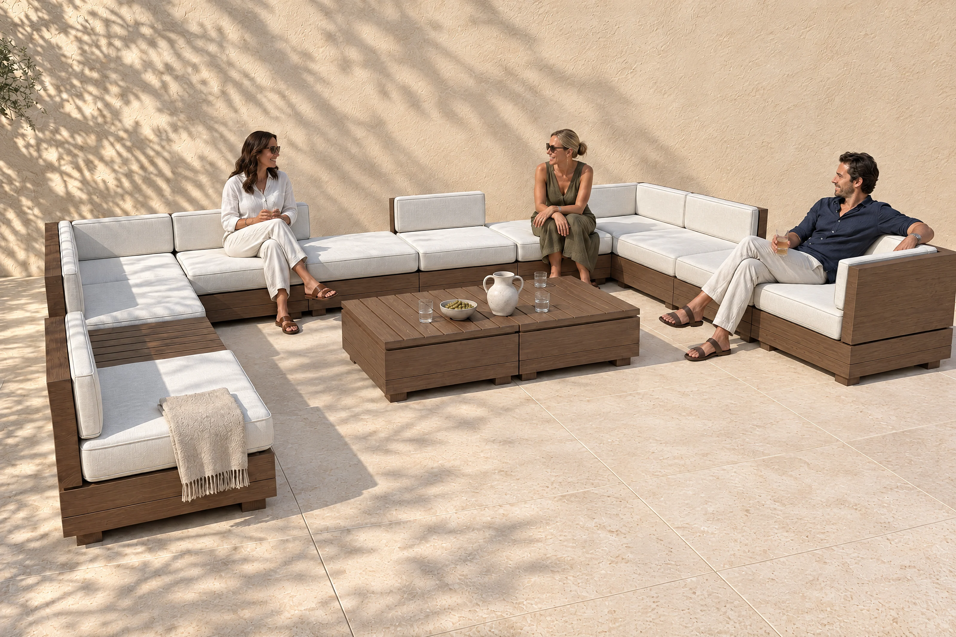 A Sama arrangement in the founder's own U on a limestone terrace, off-white cloth on walnut-coloured timber, three people talking, the shadow of an olive tree on the plaster wall behind.